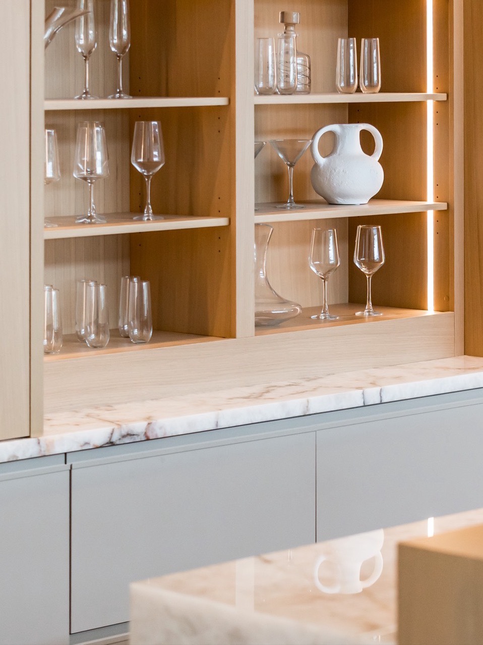 Lit white oak display shelving with glassware above a marble-topped cabinet